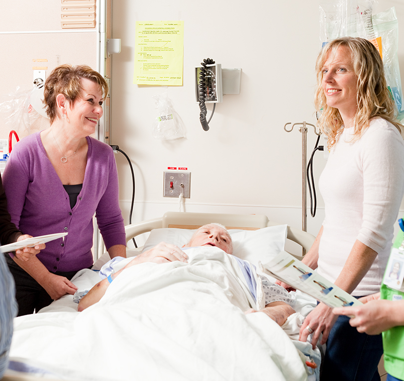 Two women clinicians stand at the bedside of a patient in an acute care setting.