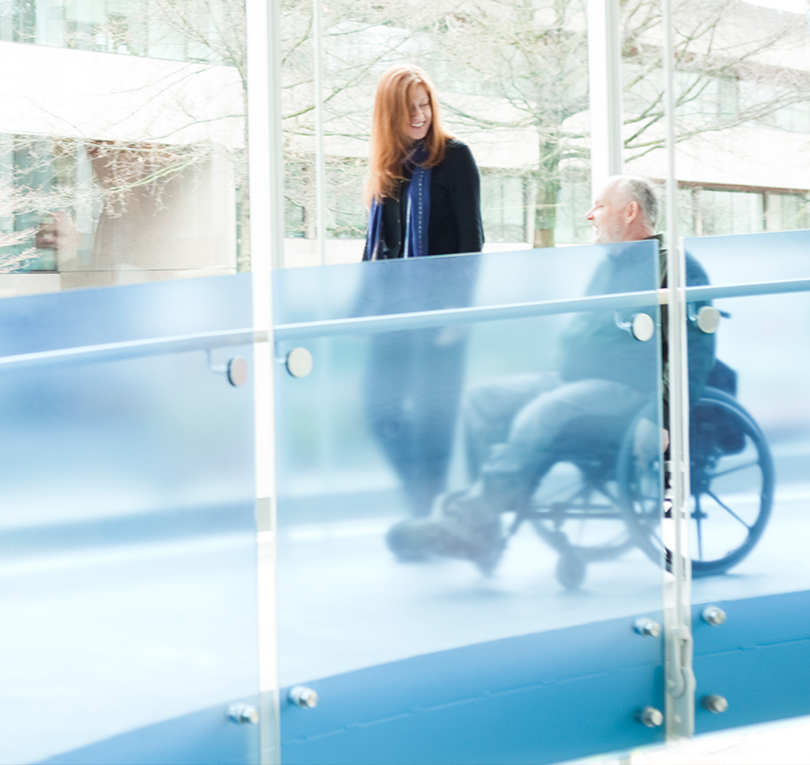 Man using a wheelchair descends a blue glass ramp alongside a walking woman