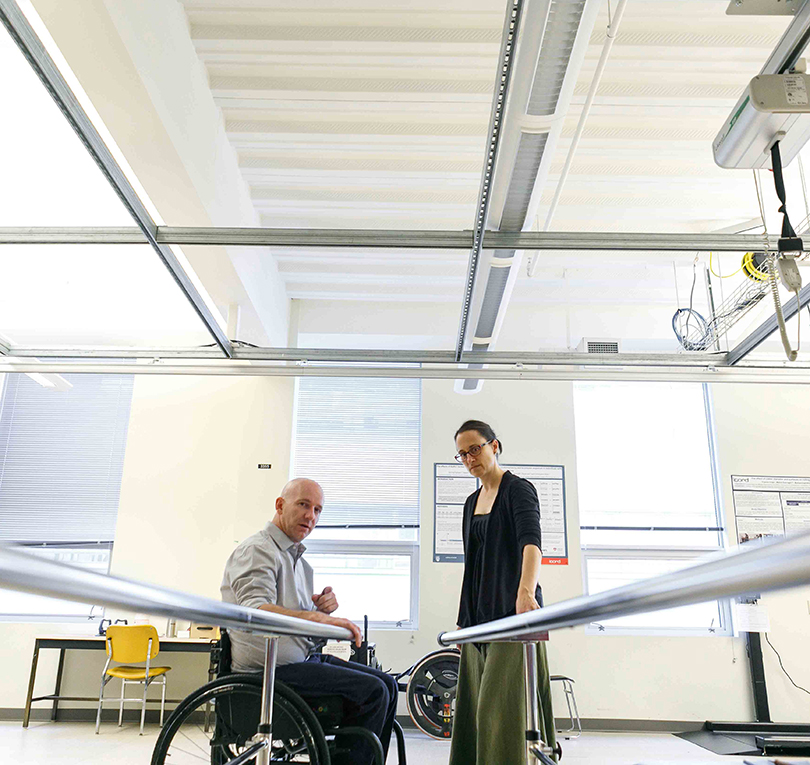 Man using a wheelchair looks at parallel metal bars with a woman standing in a rehab setting.