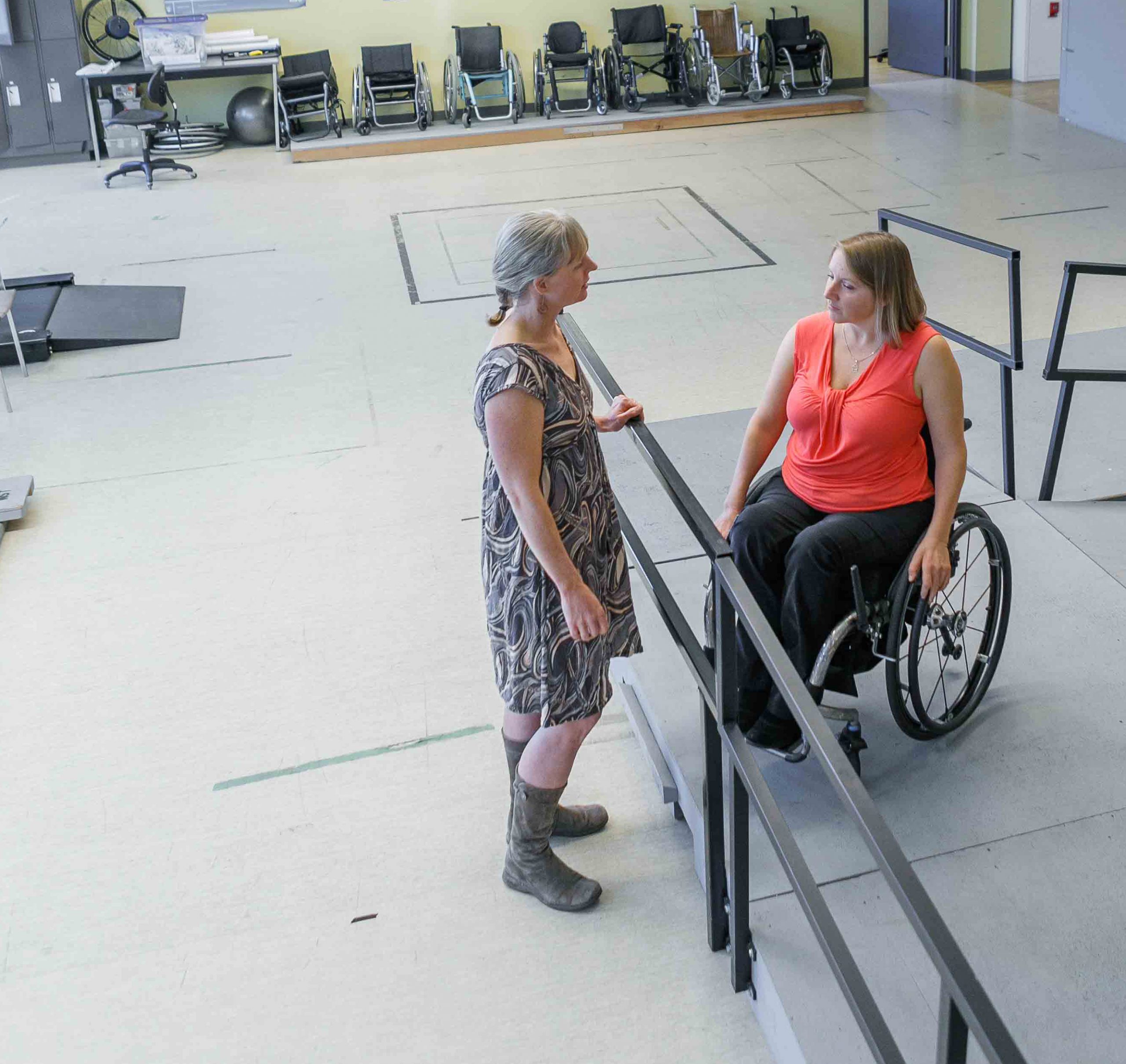 Woman using a wheelchair in an orange shirt on a ramp talks with a woman standing at a rail.