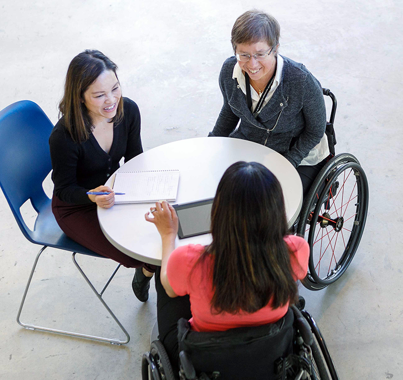 Three women using wheelchairs sit around a table in conversation.