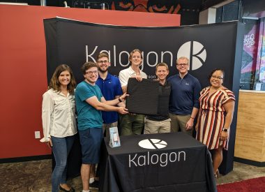 Kalogon team standing in front of Kalogon logo at trade booth